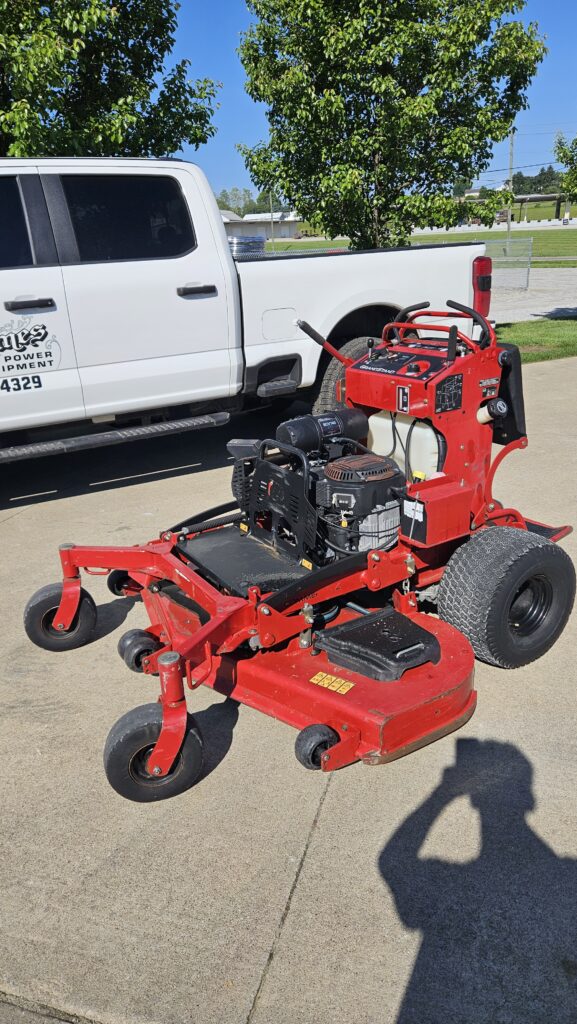 Red riding lawn mower with a gas engine parked on a concrete lot, behind a white pickup truck under green trees.