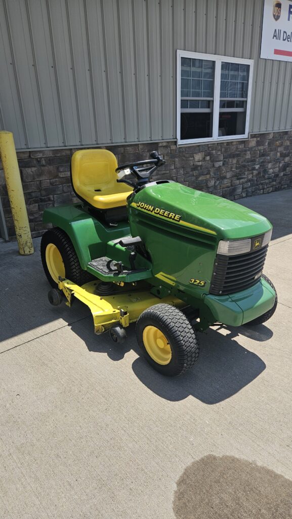 Green John Deere riding lawn mower with yellow seat parked on concrete outside a building.
