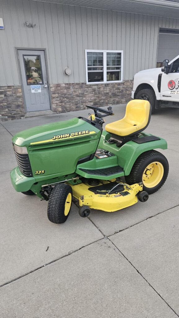 Green John Deere riding lawn mower with yellow seat parked in a concrete lot beside a gray industrial building and a white truck in the background.