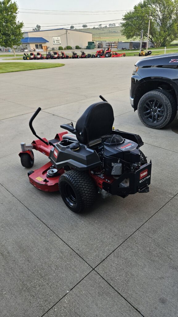 Red Toro riding lawn mower with a black seat and engine, parked on a concrete surface with a pickup truck nearby and a row of equipment in the background.
