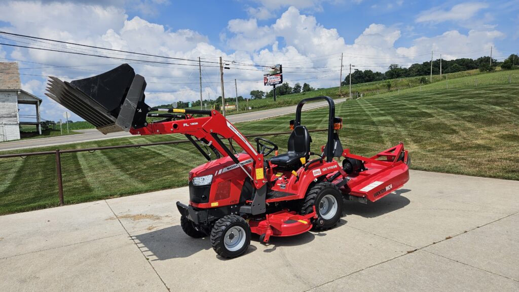 Red compact tractor with a raised front loader attached, parked on a concrete driveway with a rural field in the background.